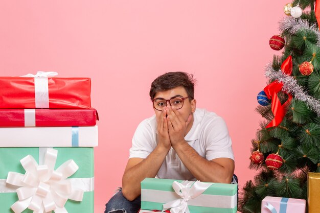 Front view of young man around presents and christmas tree on pink wall