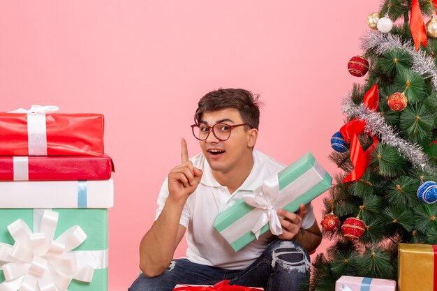 Front view of young man around presents and christmas tree on pink wall