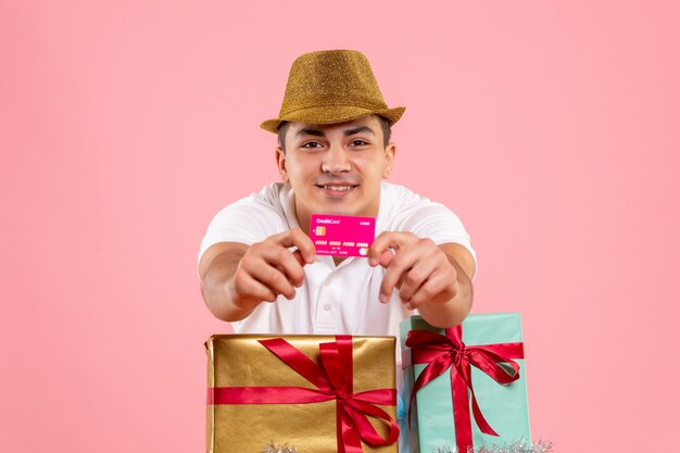 ピンクの壁に銀行カードを保持しているクリスマスプレゼントの周りの若い男の正面図