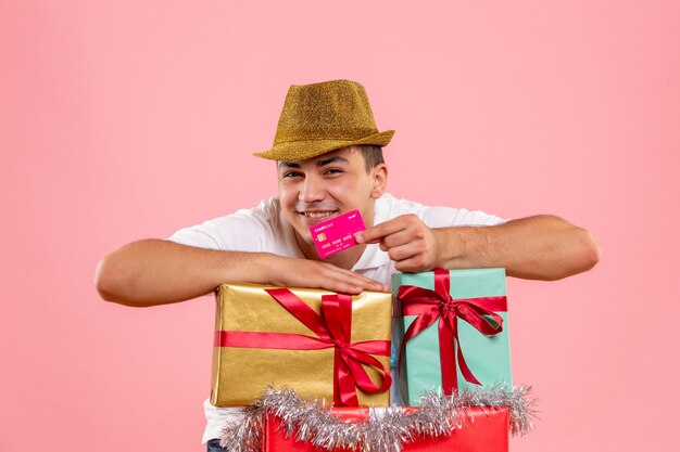 Front view of young man around xmas presents holding bank card on pink wall