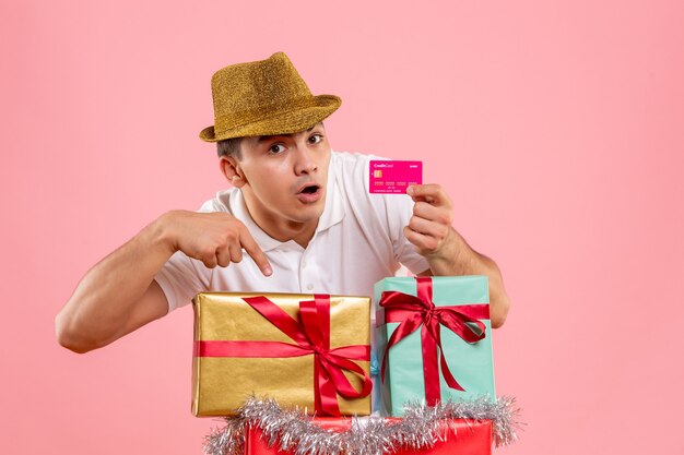 Front view of young man around xmas presents holding bank card on the pink wall