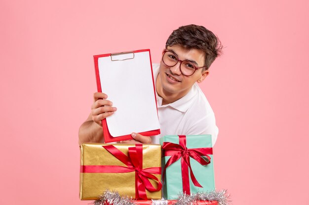 Front view of young man around xmas presents holding file note on pink wall