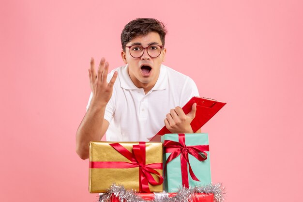 Вид спереди молодого человека вокруг рождественских подарков, держащего заметку на розовой стене