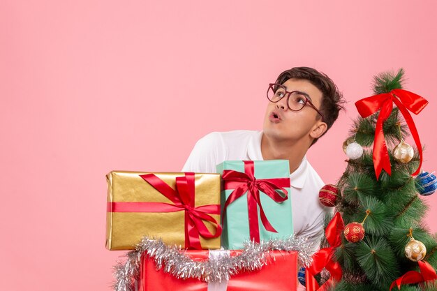 Front view of young man around xmas presents and holiday tree on pink wall