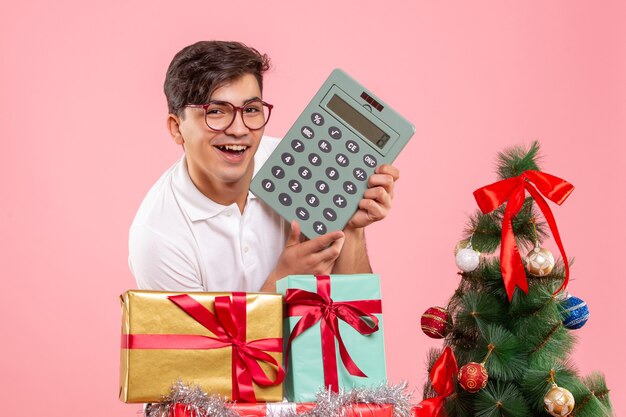 Front view of young man around xmas presents with calculator on a pink wall