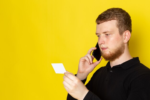 Front view of young man in black shirt talking on the phone holding white plastic card