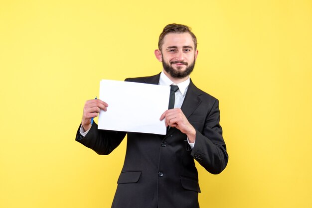 Front view of young man in black suit cheerfully smiling and holding white blank paper sheets on yellow