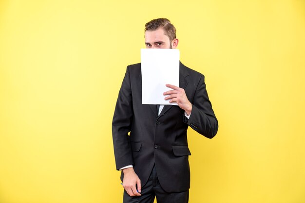 Front view of young man in black suit hiding lower part of face with blank paper on yellow wall