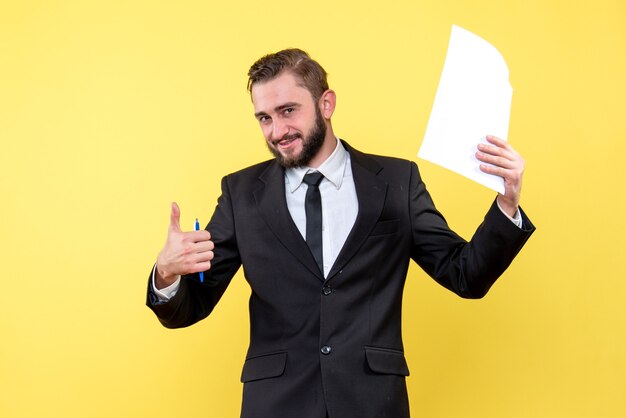 Front view of young man in black suit holding blank paper over yellow with smile thumb up with fingers excellent sign