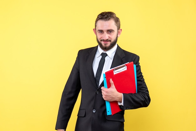 Front view of young man in black suit holding red clipboard and blue folder smiling on yellow