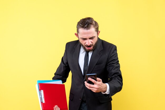 Front view of young man in black suit suffering look at a mobile phone and holding folders on yellow