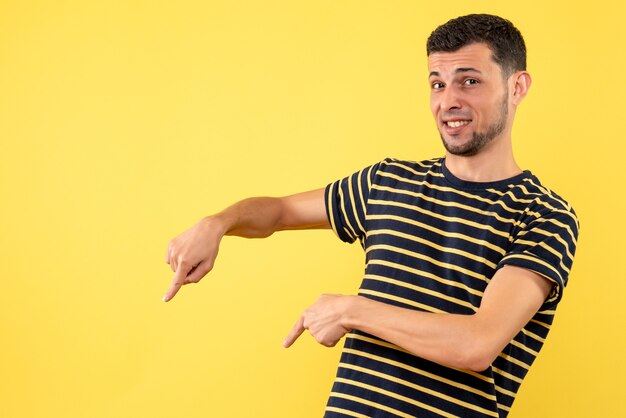 Front view young man in black and white striped shirt pointing at floor on yellow isolated background