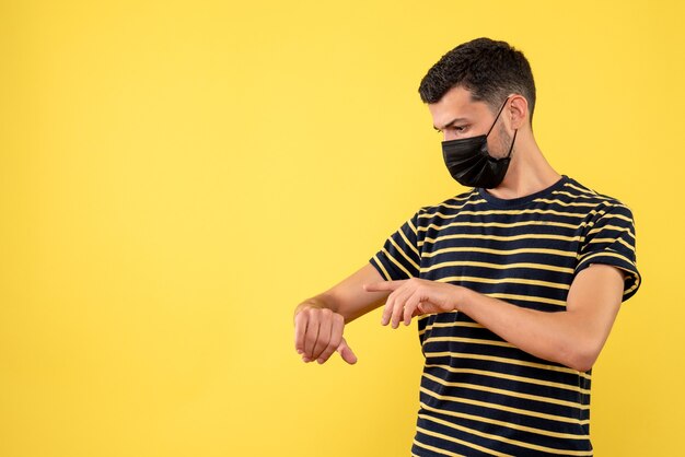 Front view young man in black and white striped t-shirt checking time on watch on yellow background