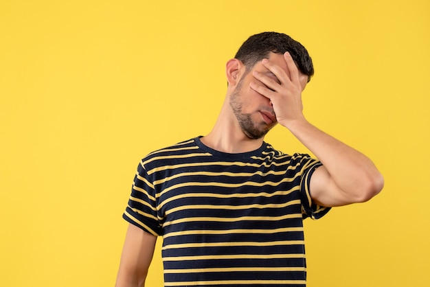 Front view young man in black and white striped t-shirt covering eyes with hand yellow isolated background