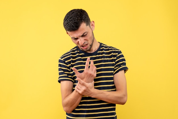 Front view young man in black and white striped t-shirt holding hand with pain on yellow isolated background