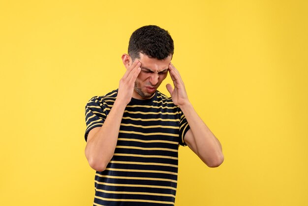 Front view young man in black and white striped t-shirt holding head with pain on yellow isolated background