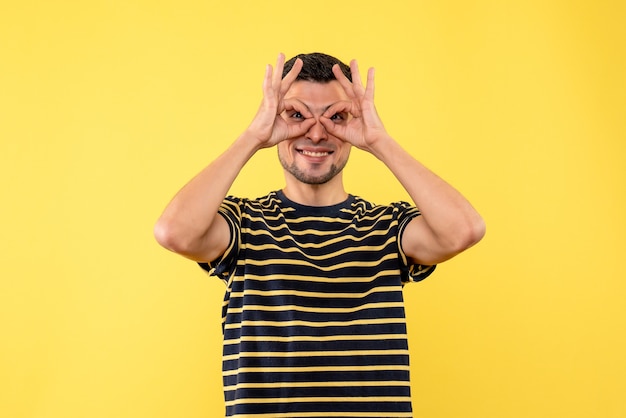 노란색 격리 된 배경에 쌍안경 손을 만드는 흑백 줄무늬 티셔츠에 전면보기 젊은 남자