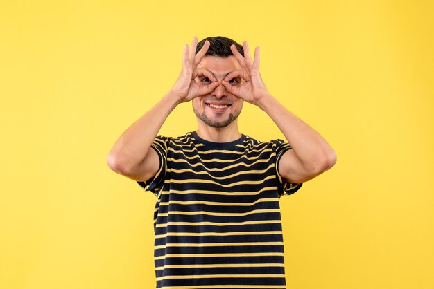 黄色の孤立した背景に双眼鏡の手を作る黒と白の縞模様のTシャツの正面図の若い男