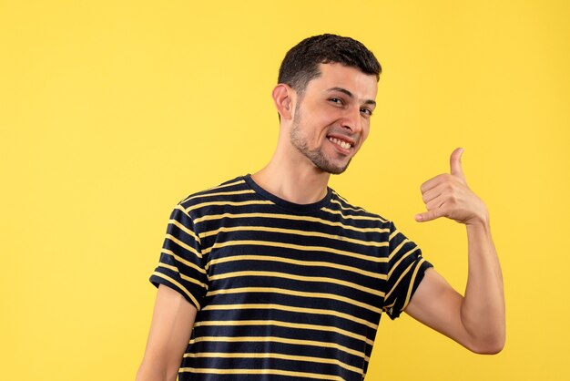 Front view young man in black and white striped t-shirt making call me sign on yellow isolated background