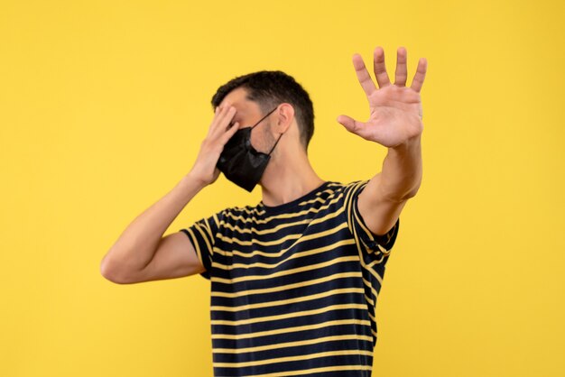 Front view young man in black and white striped t-shirt making stop sign closing eyes on yellow background