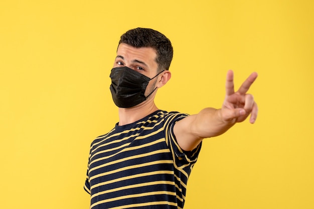 Front view young man in black and white striped t-shirt making victory sign yellow background