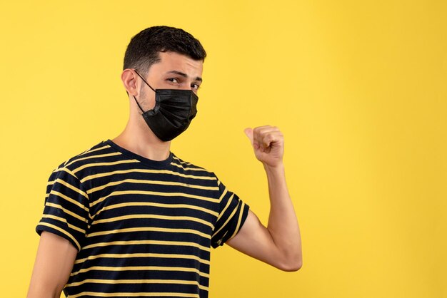 Front view young man in black and white striped t-shirt pointing with finger back on yellow background