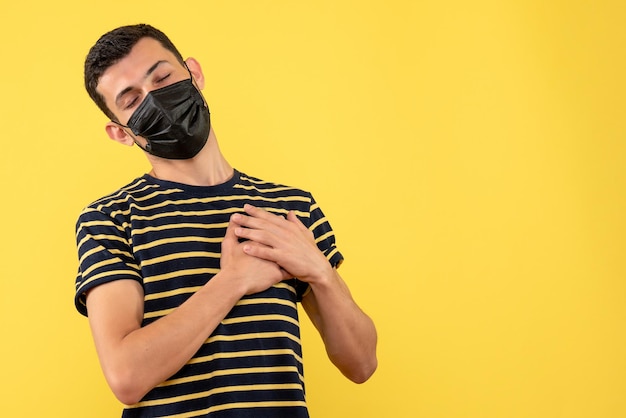 Front view young man in black and white striped t-shirt putting hands on heart on yellow isolated background