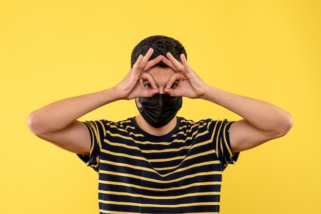 Free photo front view young man in black and white striped t-shirt putting okey sign in front of his eyes on yellow background
