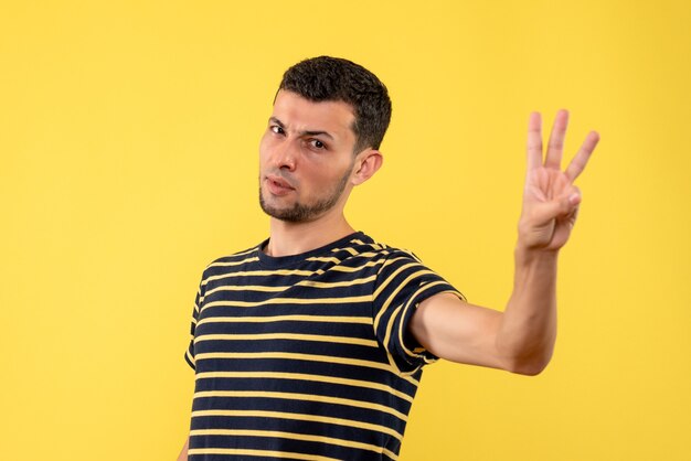 Вид спереди молодой человек в черно-белой полосатой футболке показывает три пальца на желтом изолированном фоне