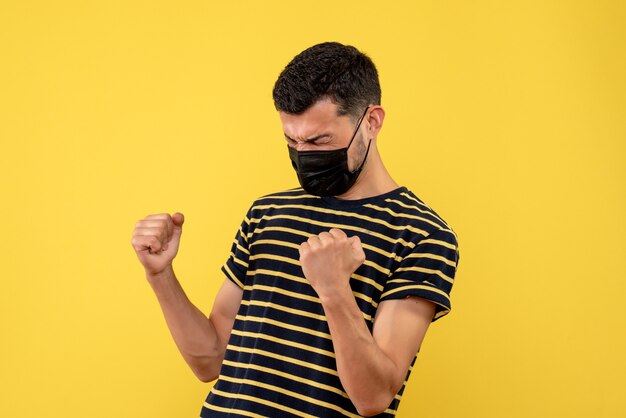 Front view young man in black and white striped t-shirt showing winning gesture on yellow isolated background