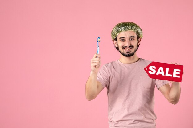 front view of young man cleaning his teeth and holding sale banner on a pink wall