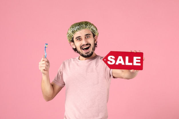 front view of young man cleaning his teeth and holding sale banner on pink wall