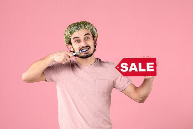 front view of young man cleaning his teeth and holding sale banner on pink wall