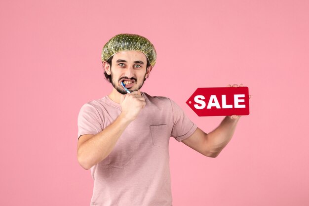 front view of young man cleaning his teeth and holding sale banner on pink wall