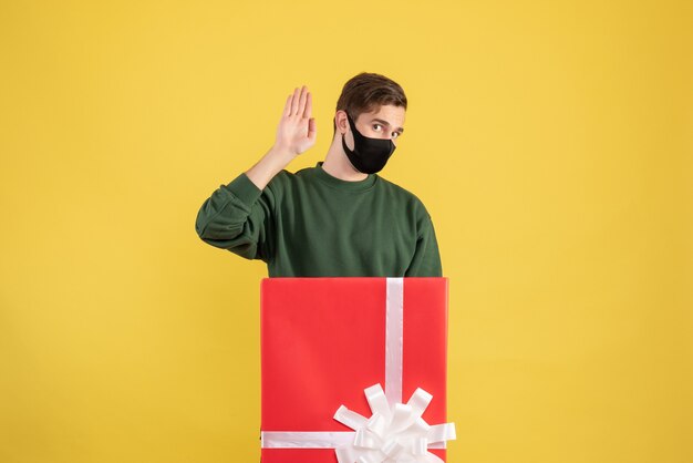 Front view young man hailing someone standing behind big giftbox on yellow 