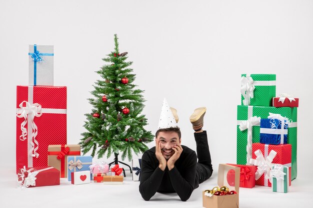 Front view of young man happily laying around holiday presents on white wall