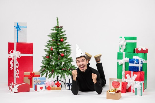 Front view of young man happily laying around holiday presents on white wall