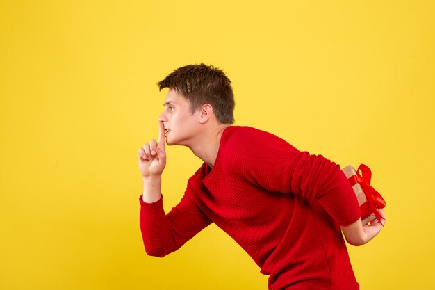 Front view of young man hiding christmas present behind his back on yellow wall