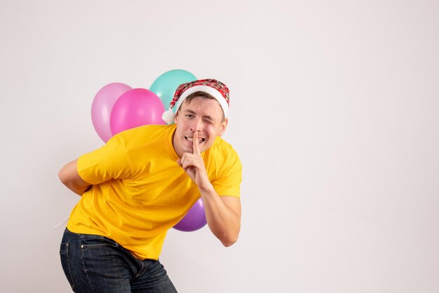 Front view of young man hiding colorful balloons behind his back on a white wall