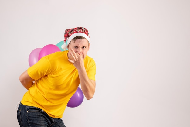 Front view of young man hiding colorful balloons behind his back on white wall