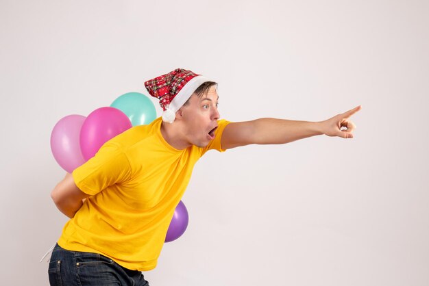Front view of young man hiding colorful balloons behind his back on white wall