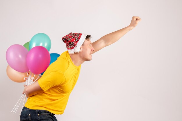 Front view of young man hiding colorful balloons behind his back on white wall