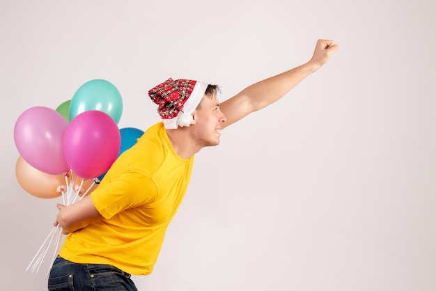 Вид спереди молодого человека, скрывающего разноцветные воздушные шары за спиной на белой стене