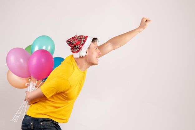 Front view of young man hiding colorful balloons behind his back on white wall