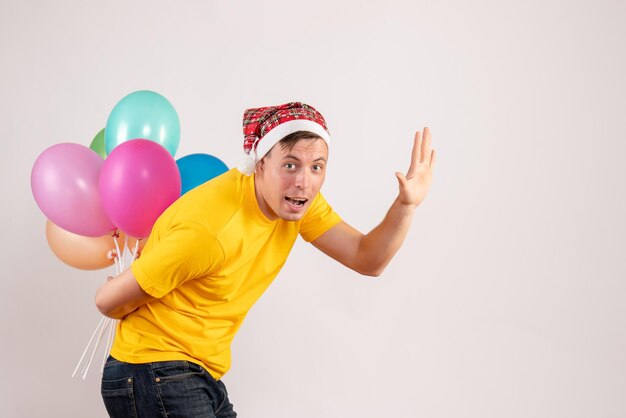 Front view of young man hiding colorful balloons behind his back on white wall