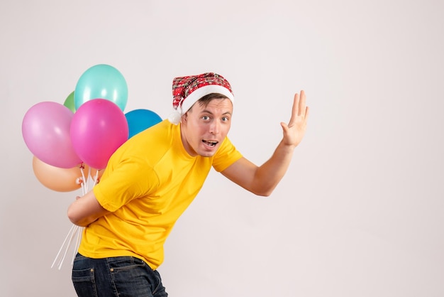 Front view of young man hiding colorful balloons behind his back on white wall