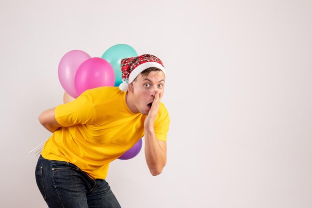 Вид спереди молодого человека, скрывающего разноцветные воздушные шары за спиной на белой стене