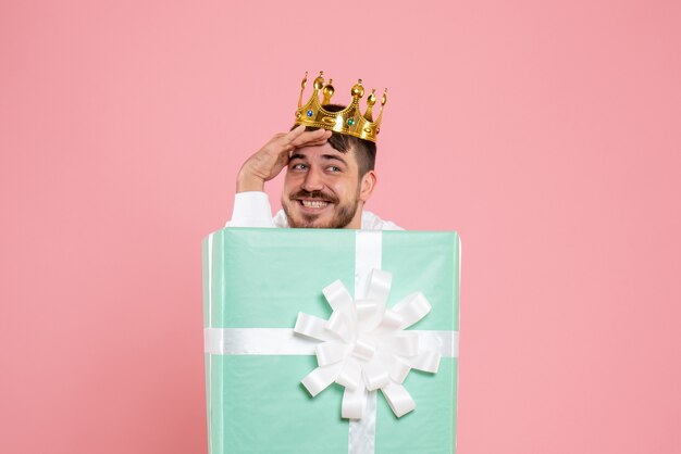 Front view of young man hiding inside present box with crown on a pink wall