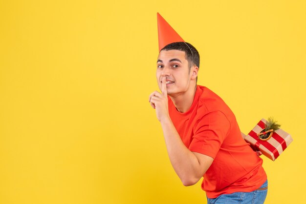 Front view of young man hiding xmas present behind his back on the yellow wall