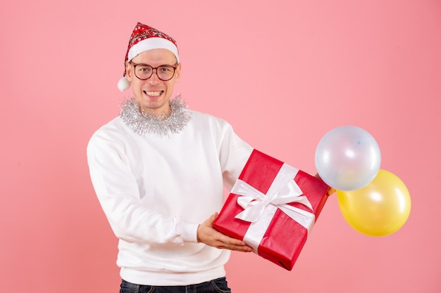 Front view of young man holding balloons and present on pink wall